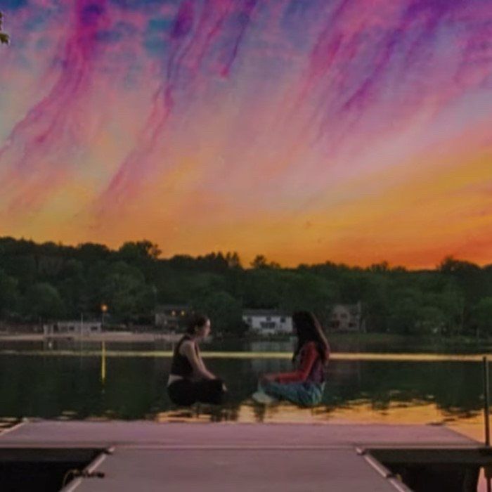 Isabel and Tara from the TV show, The Pink Opaque, sit criss-cross-applesauce, hovering a few feet above the dock on the lake at summer camp. The sky above them is all sorts of weird psychedelic colors. 
