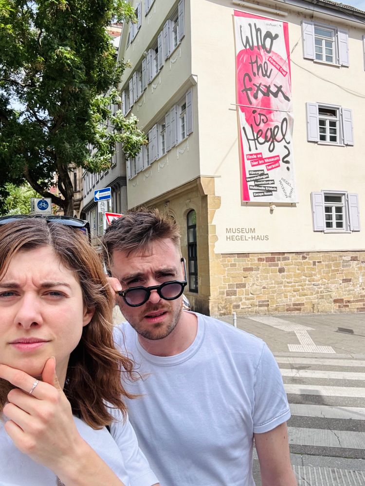 A woman and man pulling over-the-top puzzled faces outside a museum with a display saying “who the **** is Hegel?”