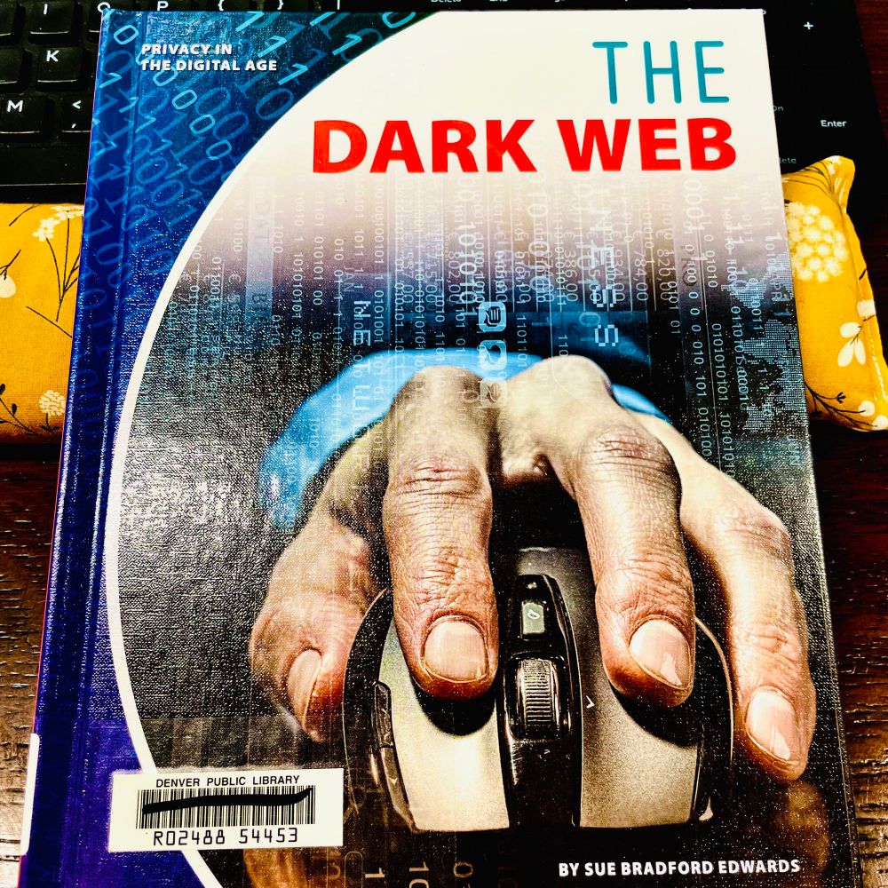A picture of a children’s book from the Denver Public Library sitting on a yellow wrist rest and a black keyboard. The cover shows a giant hand holding a computer mouse facing toward the viewer. The text says that it’s part of the “Privacy in the digital age” series. Title is “THE DARK WEB” by Sue Bradford Edwards.