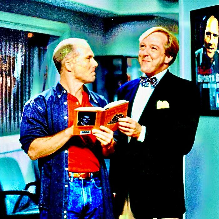 A screenshot from the TV show Frasier of Bob Bulldog Briscoe—a balding man in an oversized jean shirt with the sleeves rolled up over a red t shirt tucked into jeans—holding a book with Gil Chesterton—a prim man wearing a black blazer over khakis with a polka dot bowtie and a whimsical expression—at the KACL radio station.