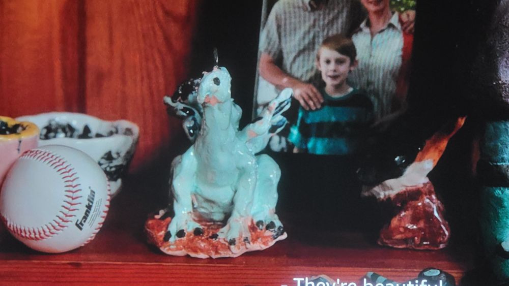 From the 2015 film, Solace, a shelf with a baseball, family photo, and a child's sculpture of a blue/green dragon. 