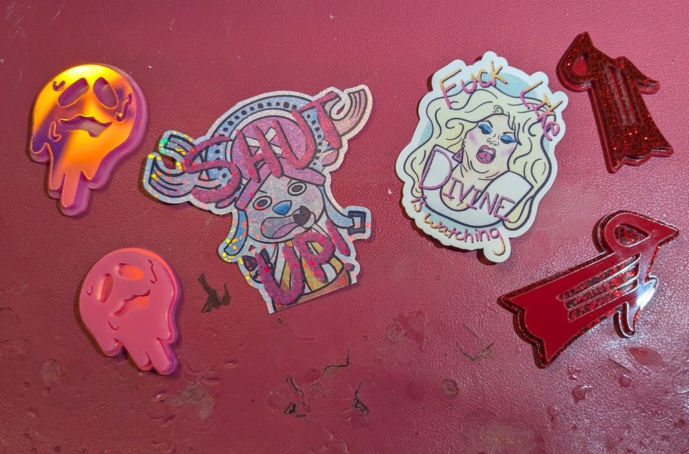 Spread featuring two Owl House guild pins (Abomination [pink and iridescent pink] and Bard [solid red and red glitter]), a holographic sticker of Tony Tony Chopper yelling SHUT UP!, and a sticker with Divine on it reading "fuck like Divine is watching" 