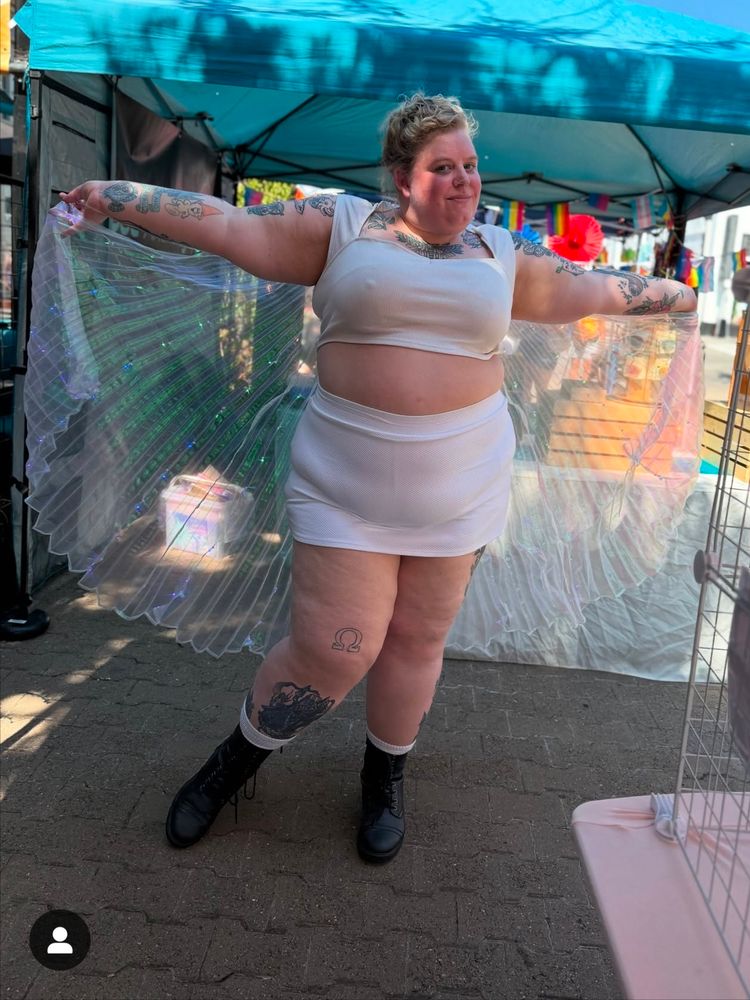 Photo of a fat blonde woman in a white crop top and short white skirt with an iridescent Isis cape on with her arms spread. 