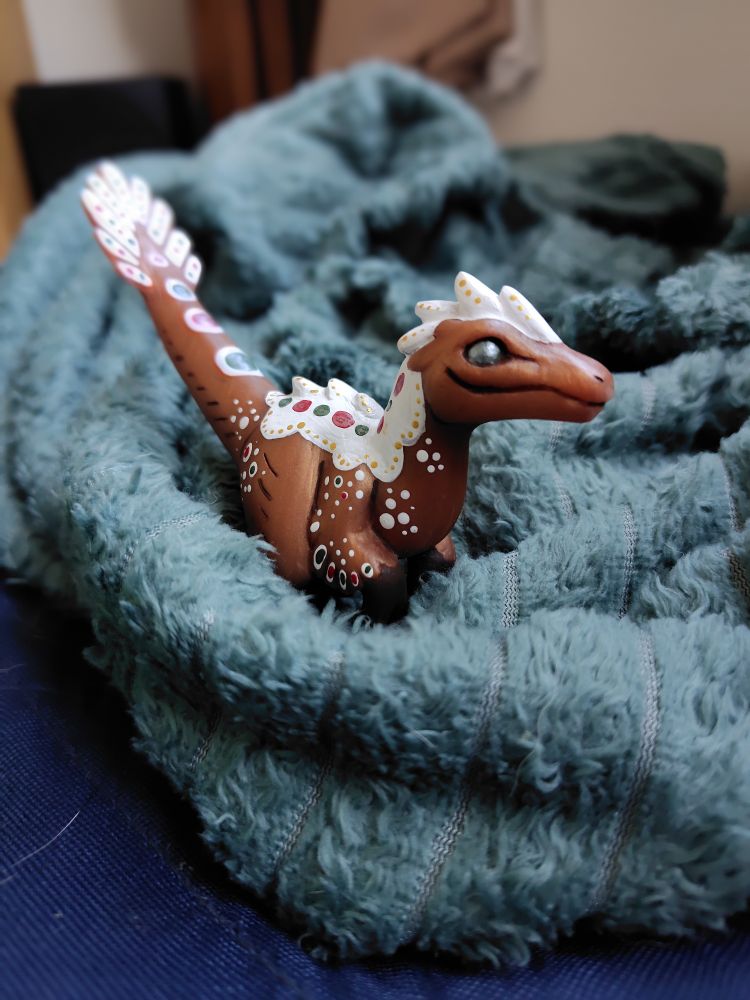 A decorative feathered velociraptor figurine made to look like it's made of gingerbread and frosting nesting in a plush green-blue blanket
