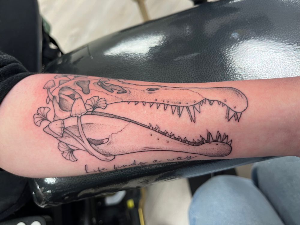a fresh tattoo on a forearm of a spinosaurus skull surrounded by ginkgoes and small mushrooms. the words “life finds a way” are written in cursive on the bottom