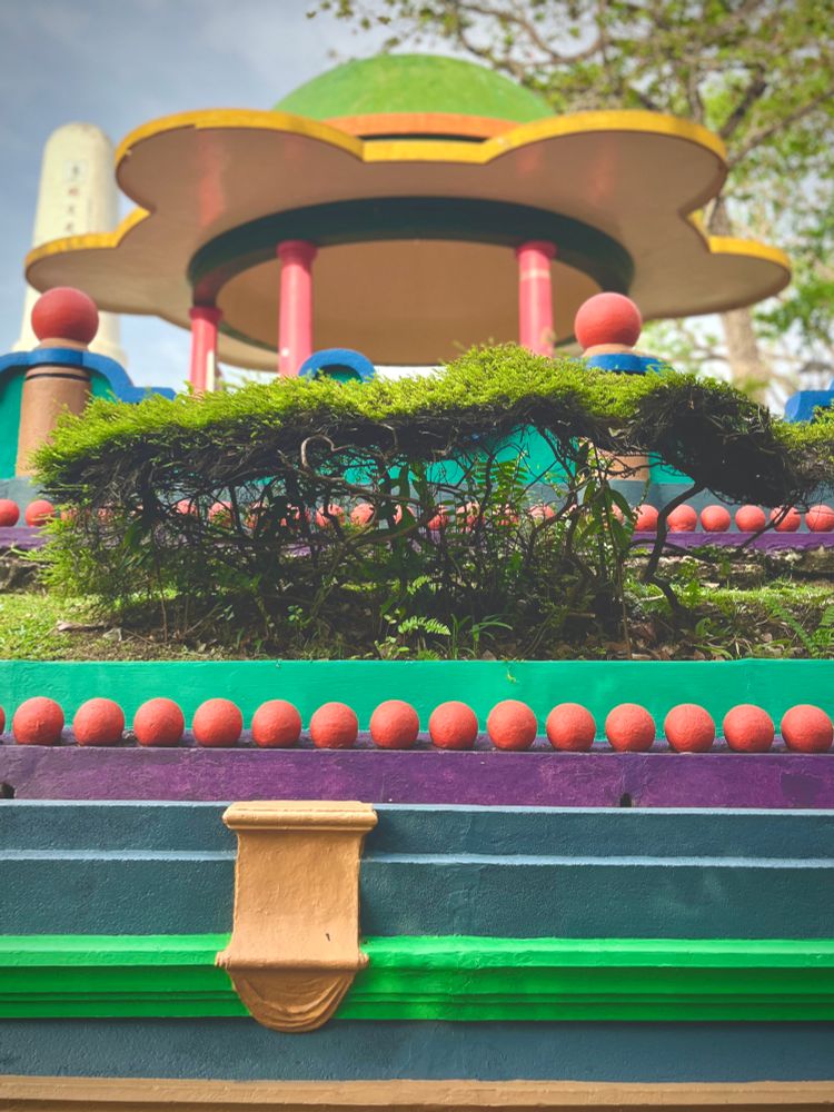 A geometricly shaped yellow pavillion. Underneath are a series of brightly coloured low walls in bold colours. Some are adorned with rows of red balls
