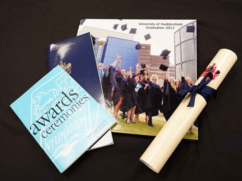 Award ceremony programmes from 1995, 2013 and 2015 laid out on a black background with a prop graduation scroll. The programmes are different shades of blue and one has a photo of graduates throwing their caps. 