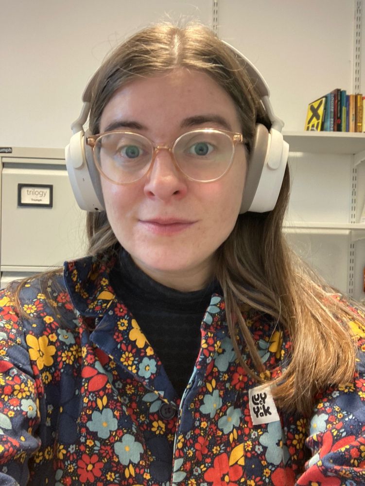 Me with my hair down, and glasses and white over-ear headphones on. I'm wearing a colourful Lucy & Yak jumpsuit with a dark roll neck top underneath. The jumpsuit has a floral pattern in blue, purple, red and yellow.