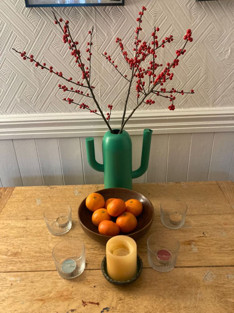 A green metal IKEA vase, shaped like a cactus, containing a bouquet of red ilex berries, standing behind a wooden fruit bowl full of oranges, which is surrounded by four tea lights in clear holders, and one larger yellow candle