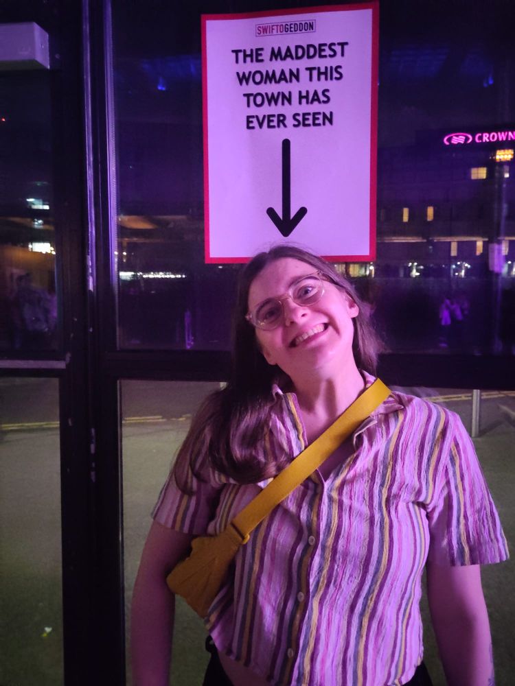 Me at a Taylor Swift club night event pulling a silly face standing under a poster with a downward pointing arrow, with the words 'The maddest woman this town has ever seen'