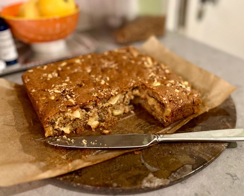 A cake on a counter. A slice has been taken out. The cake is brown with visible chunks of  apple. There are toasted walnuts on top.