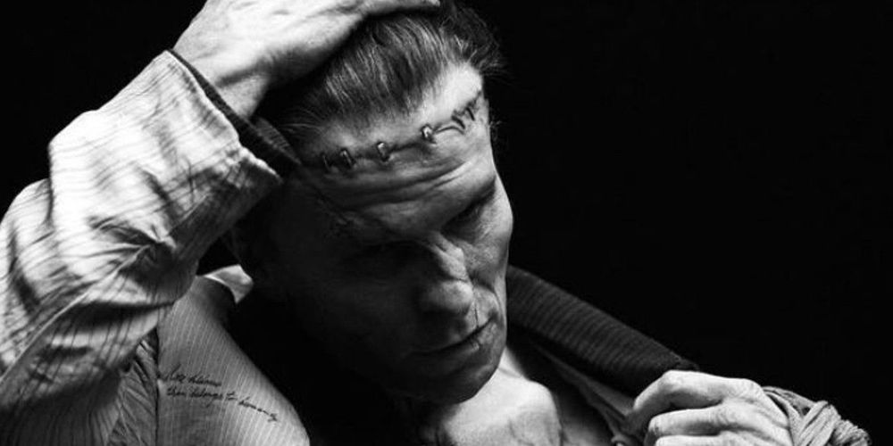 A somber production still of Christian Bale as the monster in the upcoming 2025 horror film Frankenstein. His stitched brow furrows with anguish as he rakes a hand through his hair, his expression caught between despair and defiance. The stark lighting casts deep shadows, highlighting the creature’s scars and the weight of his unnatural existence.