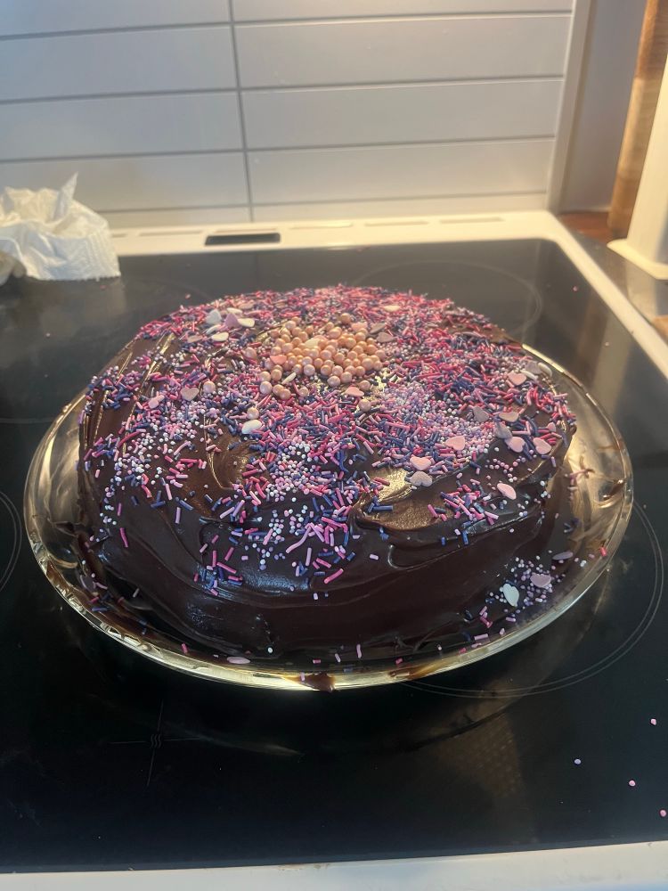 Sjokoladekake på en kjøkkenplate med enormt mye rose og lilla strø. Den sitter på en glassplate ment for kaker.