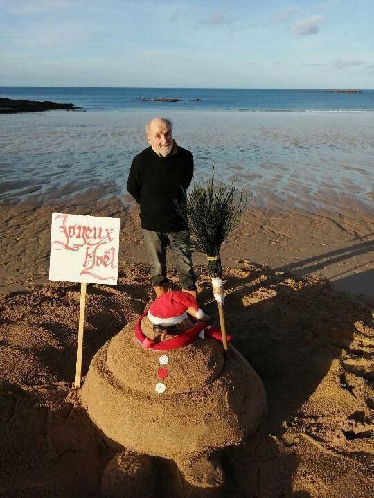 Photo d'un monsieur sur une plage derrière un bonhomme de neige fait en sable. Un bonhomme de sable donc, mais qui ressemble beaucoup à une crotte de chien un peu molle. Sauf qu'elle a un bonnet de père noël, des boutons rouges et blans, une écharpe rouge autour du... Cou ? Et un balai planté dans le dernier repli du bas. À gauche, il y a aussi un panneau plan avec écrit en rouge dessus, en lettres gothiques : Joyeux Noël.