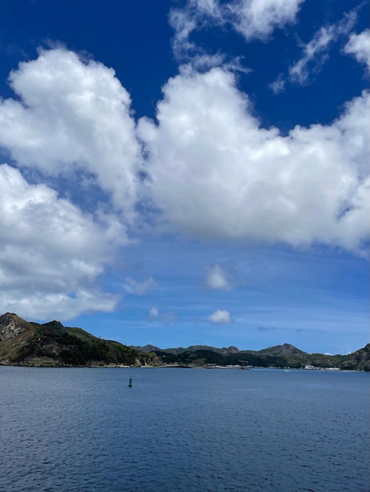 海からのぞむ島の写真。(フェリーから撮っている)青空に白い雲が浮かに、島は手前に港の入江がある。手前は深い青い色の海。