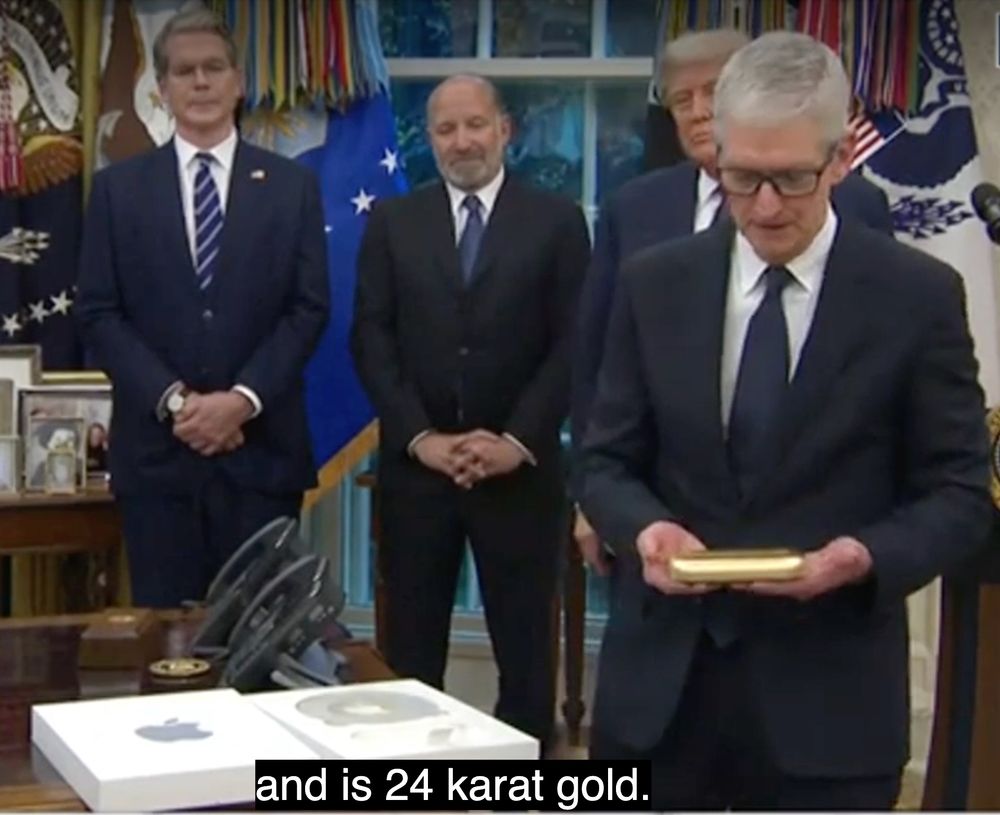 Apple CEO Tim Cook in the Oval Office preparing to hand over a 24 karat gold bribe to Donald Trump. Behind him, Donald Trump and other men in suits look on. On the desk in the foreground are white boxes with the Apple logo.
