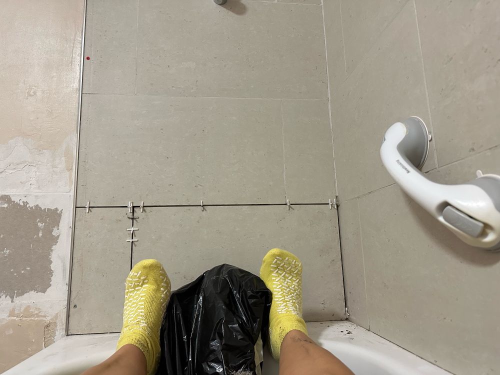 Stone coloured tiles in bathroom at the end of the bath. With two feet with yellow hospital socks as they are great for grip. Having fallen in bath this year I am being extra careful so sat in bath to do this little job. Had help to get in so need a little rest before I get out… hence am laying down!