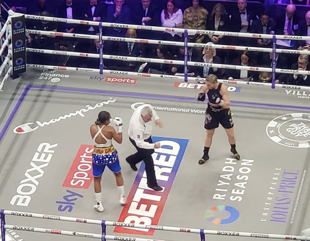 Women's world title unification boxing - the referee signals for Natasha Jonas and Lauren Price to resume fighting.
Jonas is in Blue shorts with a white vest and gloves.
Prices is all in black. 