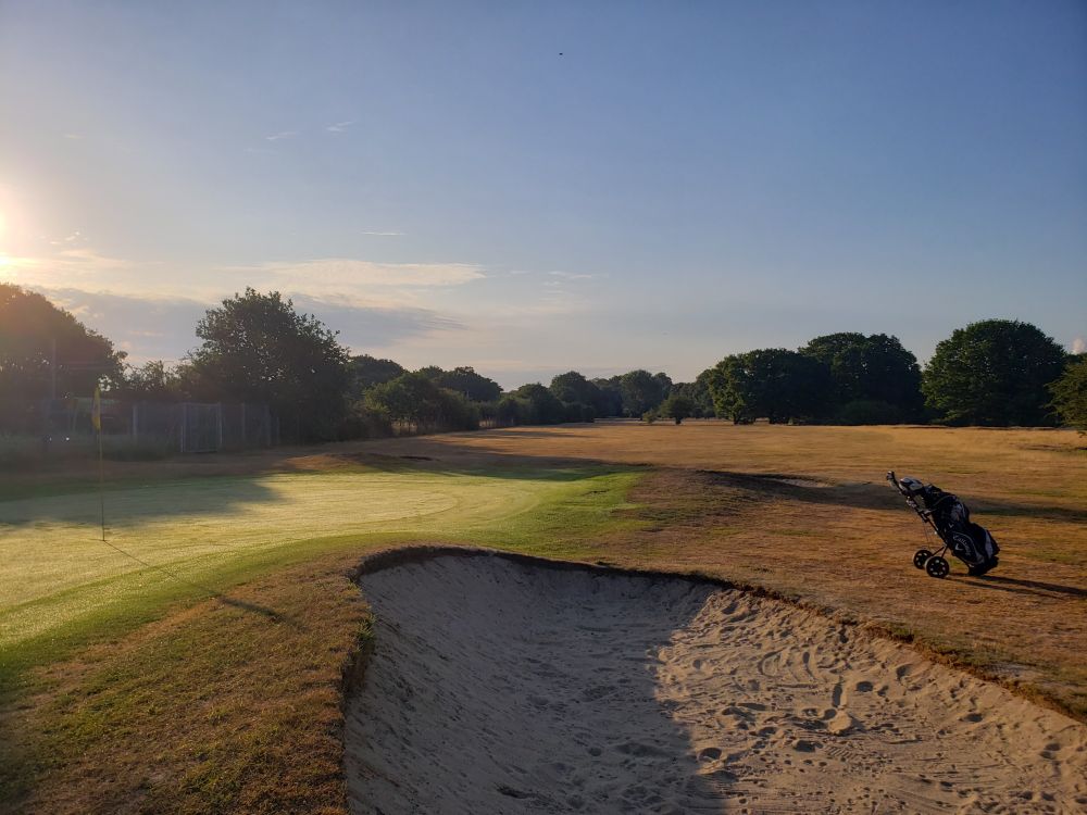 The foreground is the 9th green of a golf course on the left with the flag on the left edge of the image. At the bottom is a messy bunker, with a golf bag on a trolley to the left.
The yellow-brown-green fairway stretches into the distance with lots of small trees.
The top half of the image is a bright, almost pristinely clear blue sky, with the glare of the sun just peaking in from the left, still low in the sky.