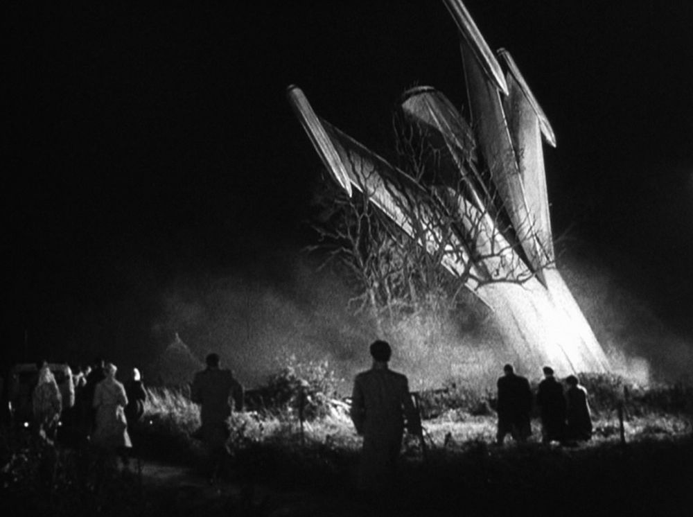 A b+w still from the 1955 horror/sf movie The Quatermass Xperiment. A rocket has crash landed and is buried nose first in the ground. The tail, with large stylised fins, is set dramatically in front of several onlookers. 