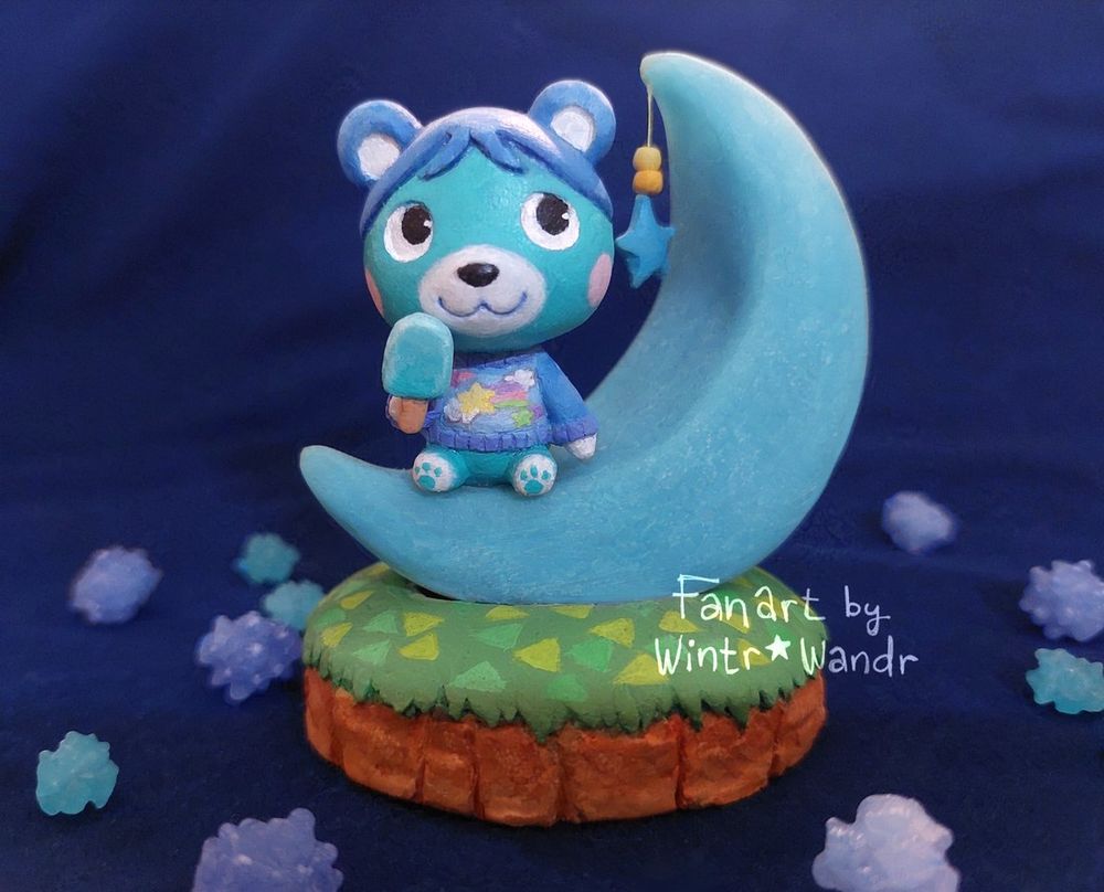A photo of a small sculpture of Bluebear from the Animal Crossing series. She's sitting on a blue crescent-moon Chair and holding a frozen treat. Surrounding the sculpture are blue konpeito candies scattered about.