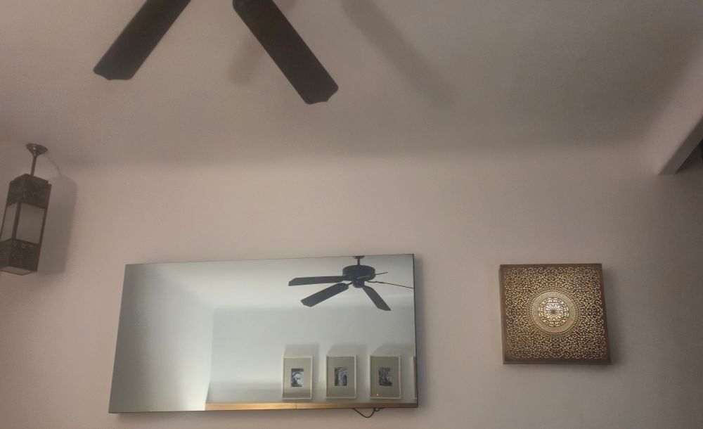 A mirror on a wall with a ceiling fan and the opposite wall reflected in it. A small electrical wire only just visible at the bottom corner of the mirror. 