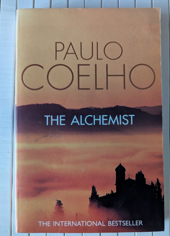 Cover of book by author Paulo Coelho. The Alchemist.  An orange coloured image with a building looming at the bottom out of the mist and hills in the background. 