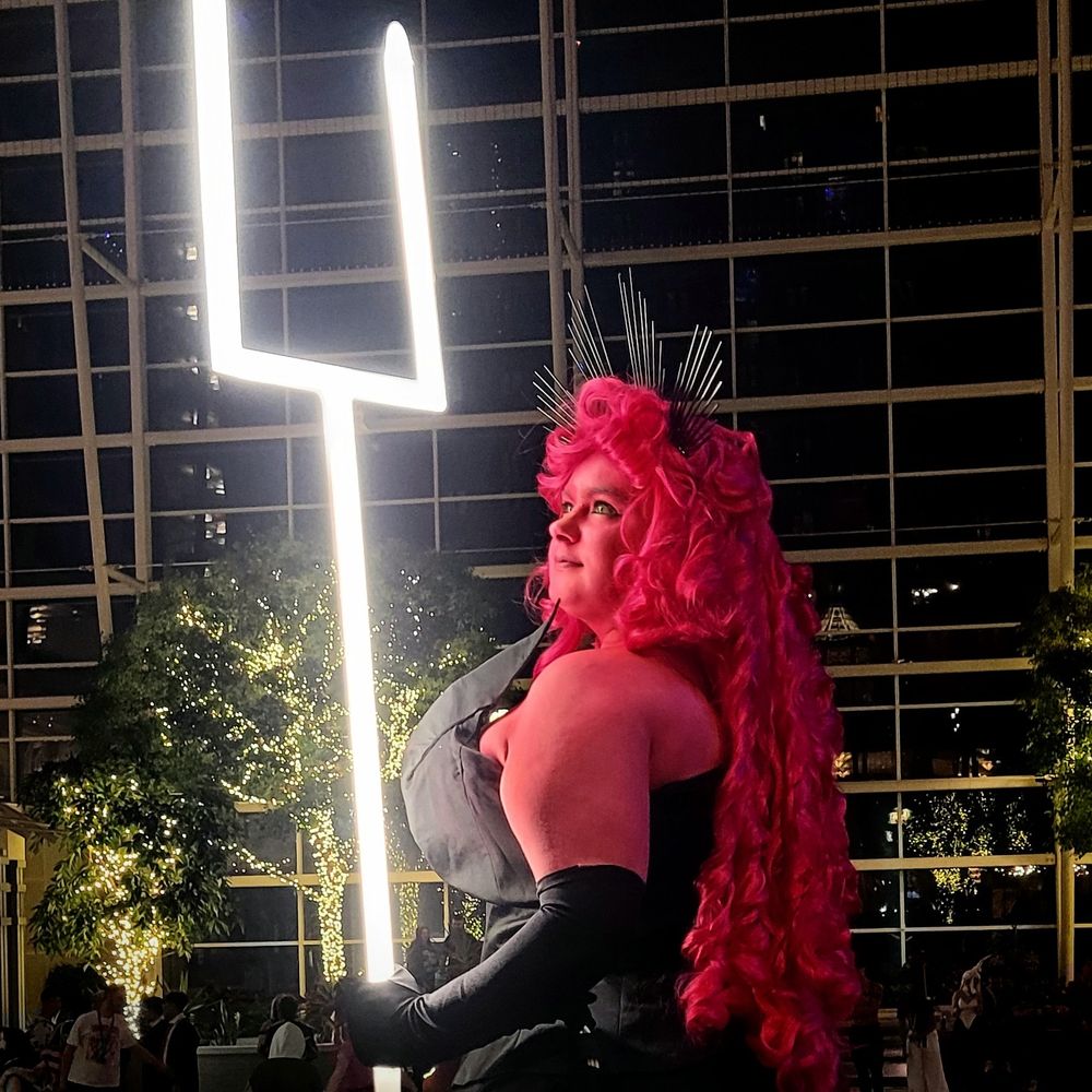 Cosplayer dressed as the character Persephone from the webcomic Lore Olympus, with pink body paint, pink wig, black dress and gloves, and a glowing bident