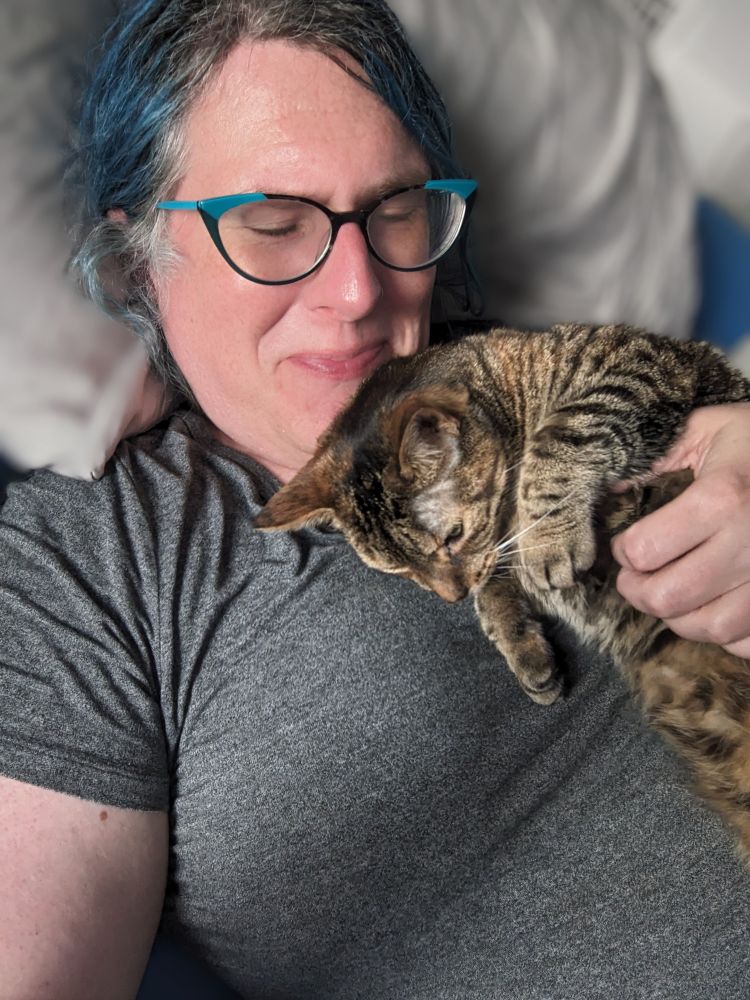 A white trans woman cuddling a tabby cat lying across her chest 