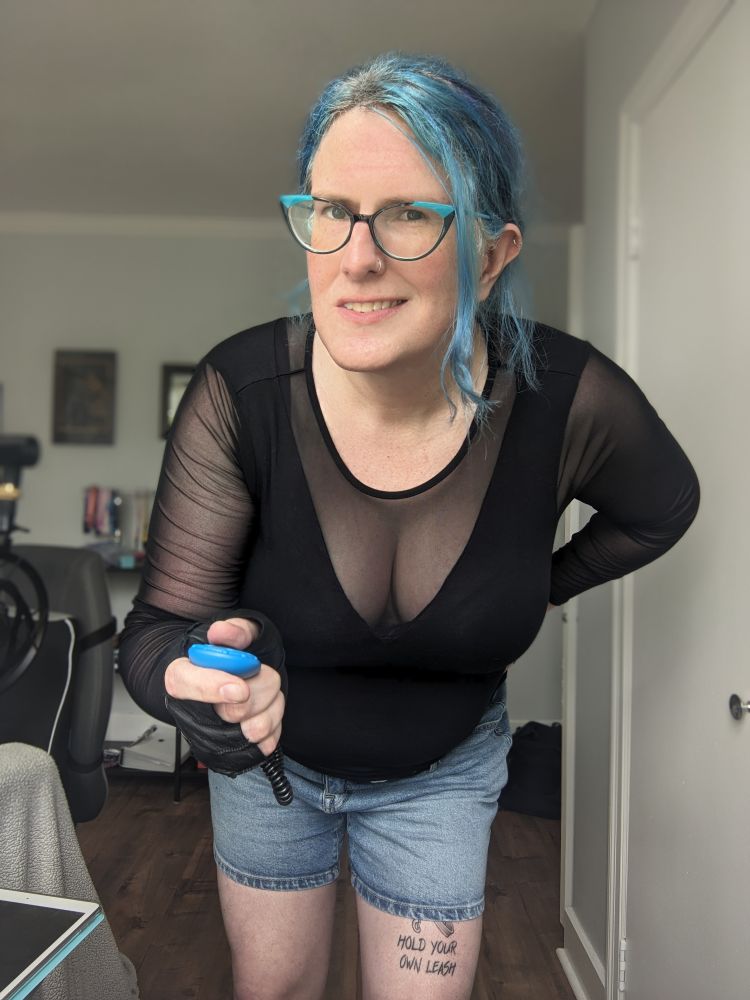 Two photos of a white trans woman with blue hair in denim shorts and a black top with mesh sleeves and a low mesh v neck, as well a fingerless leather gloves. She's leaning forward, smirking, and holding a pet-training clicker 