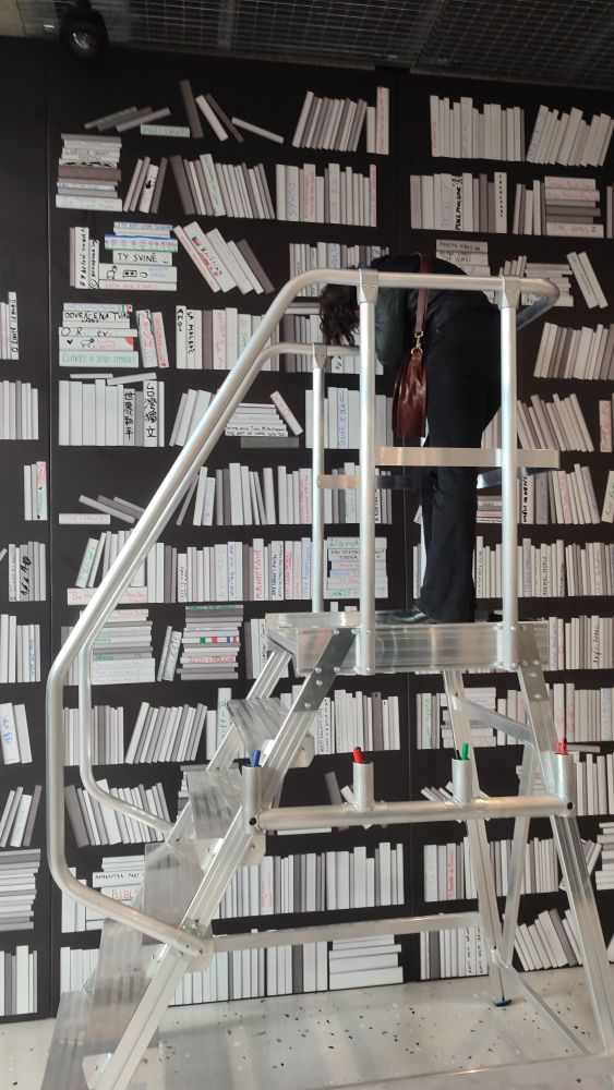 Eine riesige unechte Bücherwand mit lauter weißen Buchrücken, davor eine Leiter, darauf eine Person die gerade einen der Buchrücken mit einem Titel beschriftet.