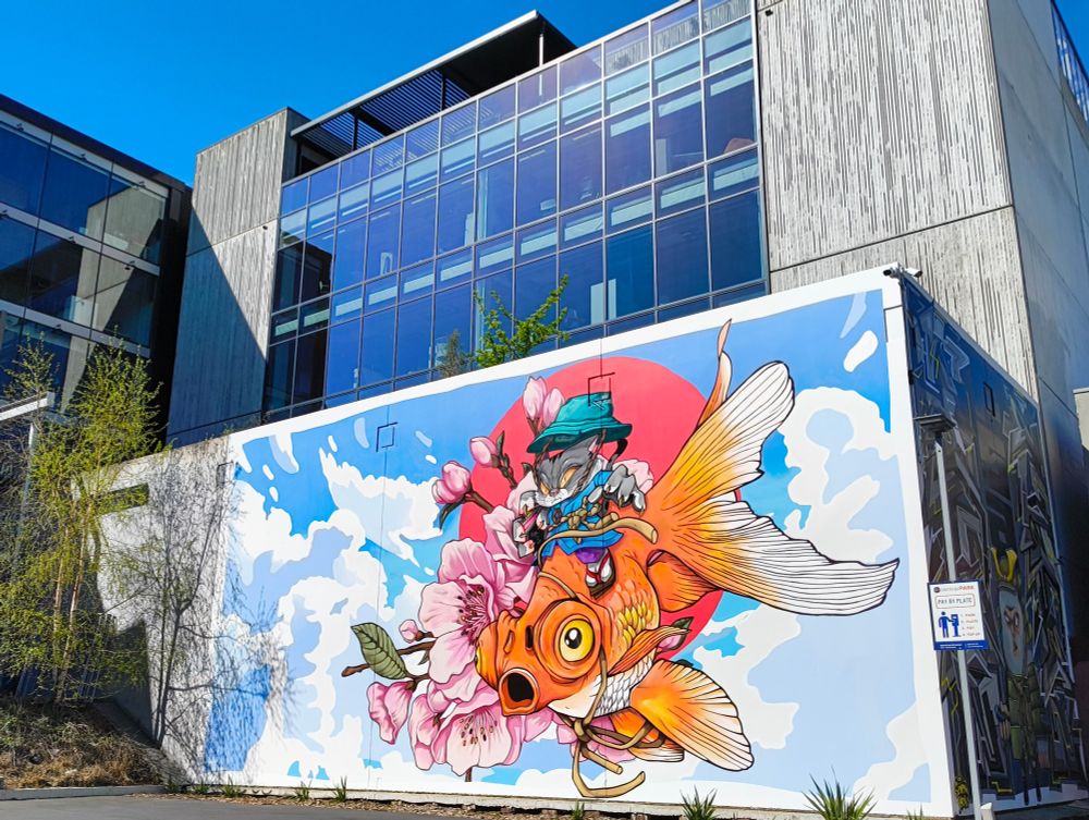 A vibrant mural by ysek7 of a cat riding a goldfish with cherry blossoms, a cloudy sky and red sun behind them