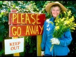 Katherine Hepburn holding a bunch of flowers smiling at the camera, stood next to two signs reading "Please Go Away" and "Keep Out"