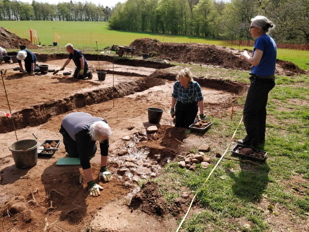 Four WallCAP volunteer excavators are digging in the trench, exposing archaeological features in a red-brown sandy soil. Dig director Jane is speaking to them from the side of the trench.