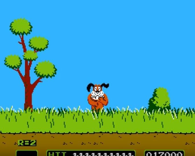 Capture d'écran d'un jeu vidéo: un paysage en 2D, pixellisé en couleurs. En premier plan un parterre de terre marron, une couche d'herbes hautes vertes au dessus. En second plan, un arbre sur la partie gauche, avec 5 touffes de feuilles au bout de ses 5 braches. Un buisson sur la partie droite. Au mileu un chien cartonnesque brun au museau blanc et aux oreilles noires, il se tient debout, surgissant des herbes hautes. son expression est moqueuse, les yeux plissés et la patte devant la bouche. 