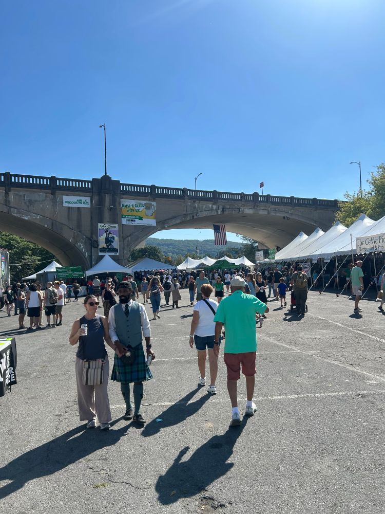 The thoroughfare outside the Grand Pavilion of Celtic Classic. Tents and crowds gather under an impressive overpass. People in kilts mill to and fro. The sky contains not a single cloud.