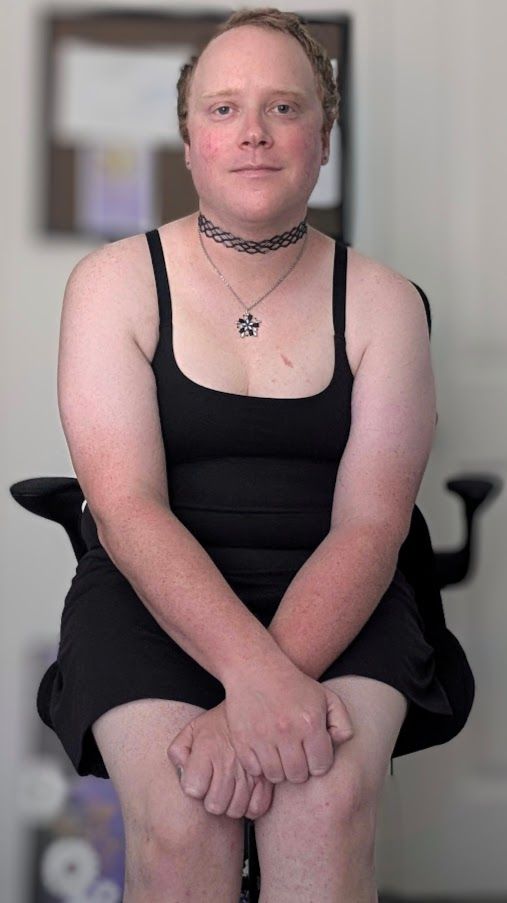 Me sitting down staring at the camera wearing a black dress that is blending into my black chair. My arms are crossed in front of me and I'm wearing a gray choker and a gray and purple pendant.