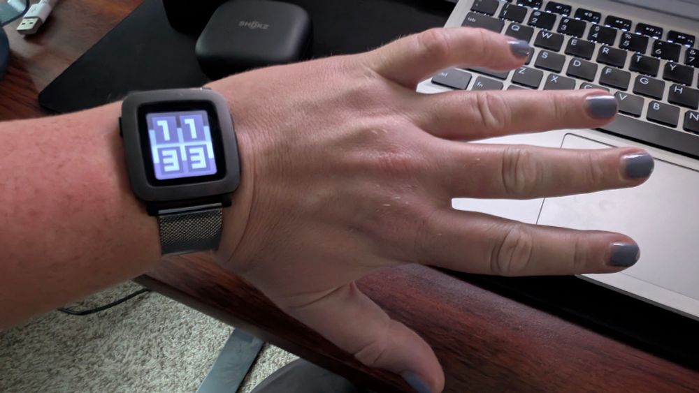 A picture of my hand with a square watch with a color eink screen that shows the time with an ace flag behind it. My nails are painted lavender, just so you know.