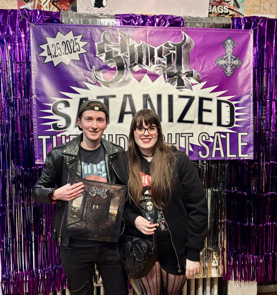 A picture of myself and my wife posing infront of a poster that says SATANIZED MIDNIGHT SALE. I am on the left and she is on the right. I am holding the new Ghost record.