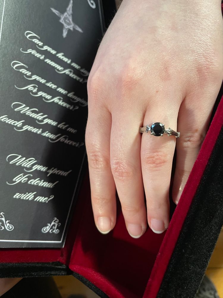 my fiancée showing her engagement ring inside the coffin box I put it in. Beside her hand is the lyrics to 'life eternal' by the band Ghost adapted as a proposal. Her engagement ring is sterling silver with a Black Swarovski gem inset. 