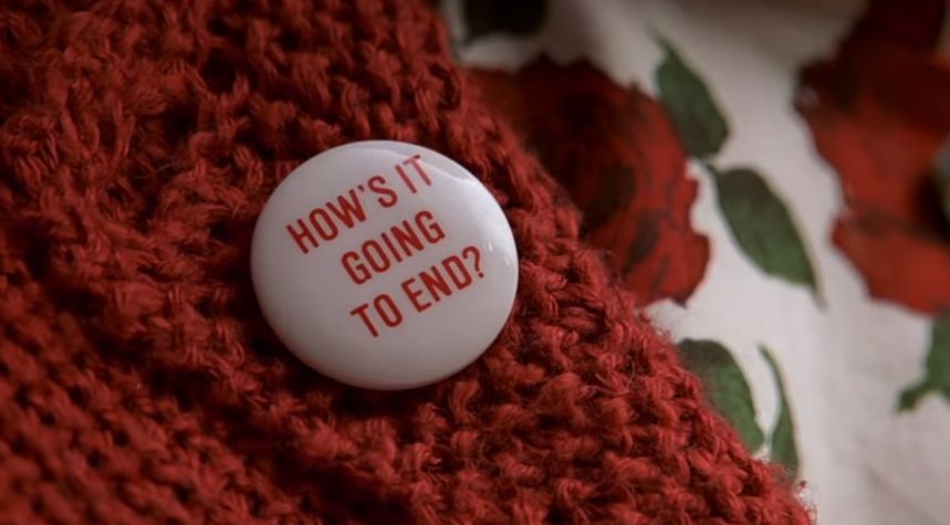 A pin badge on a red cardigan says “how’s it going to end?”
