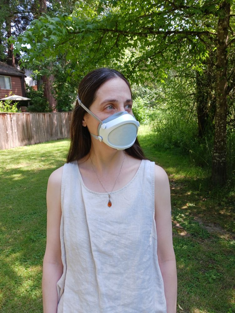 Me, in a Flo Mask in a narture-y public space. I am wearing a linen sleeveless top and an amber necklace, with a neutral expression on my face.