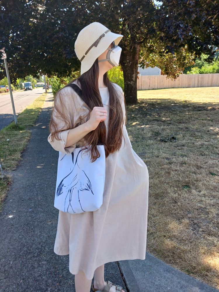 A photo of me in a sun hat, sunglasses, and a Flo Mask. I am wearing a flax-colored dress and using a tote bag featuring one of my crane drawings (available in my shop!).