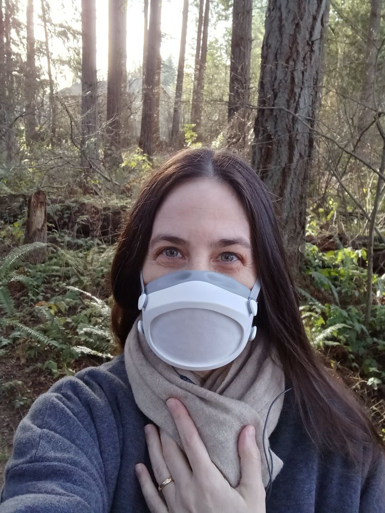 A photo of me in a scarf, coat, and Flo Mask. I am on a public path in a wooded area. 