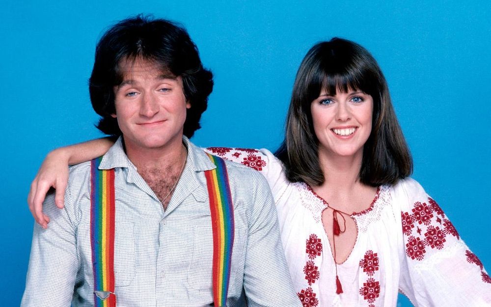 The doctor appears to be a white man with brown hair. He wears a white shirt with wide collar and vibrant rainbow suspenders. His companion is a white woman with shoulder length brown hair. She is wearing a white blouse with red flowers in a design on her arms and front.