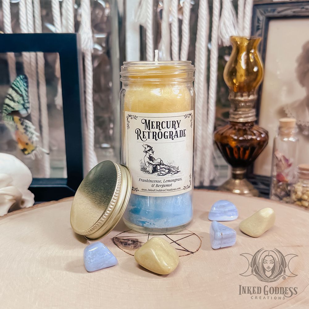 A narrow jar candle is on a wood slice with small tumbled blue lace agate and yellow aventurine gemstones around it. The candle is yellow at the top and blue at the bottom. The jar has a screw-on metal top, which is leaned against the candle jar. The jar's label reads "Mercury Retrograde" with a list of ingredients and their magickal properties. This is an exclusive product from Inked Goddess Creations.