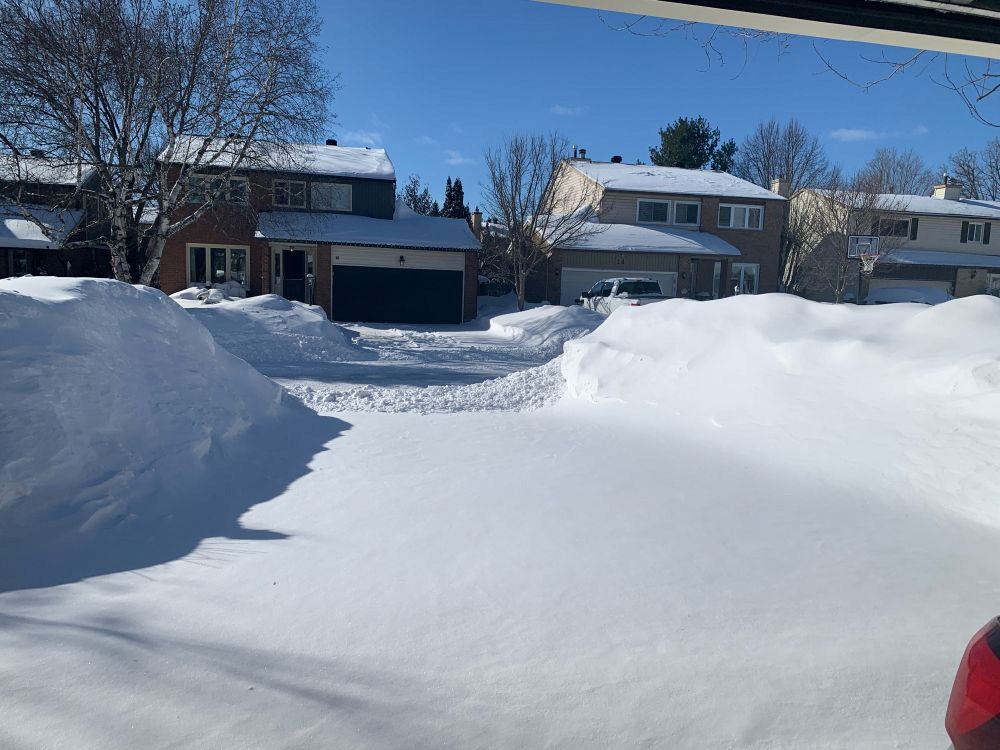 Driveway covered in snow with the end covered in plowed up ice and hardened snow. The banks on either side are almost a couple of metres high.