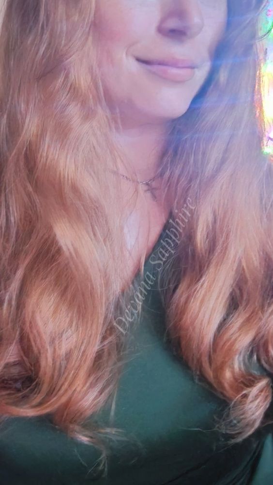 Selfie showing my long red hair falling in soft curls down my shoulders. I'm wearing a silver cross necklace and a green dress.