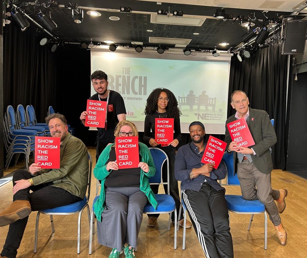 Six people (three sitting and three standing) holding red 'Show Racism the Red Card' signs in the rehearsal room at Gala Durham in front of a screen that features the lead image for The Bench by Jeff Brown. The group includes playwright Jeff Brown, producer Carole Wears and the team from Show Racism the Red Card.