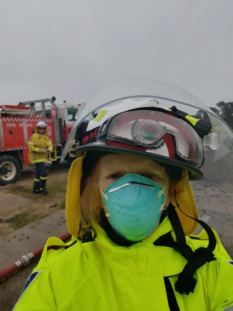 Susie, wearing firefighter uniform and helmet. Also wearing an N95 mask. In the background is a RFS truck and another Firefighter.